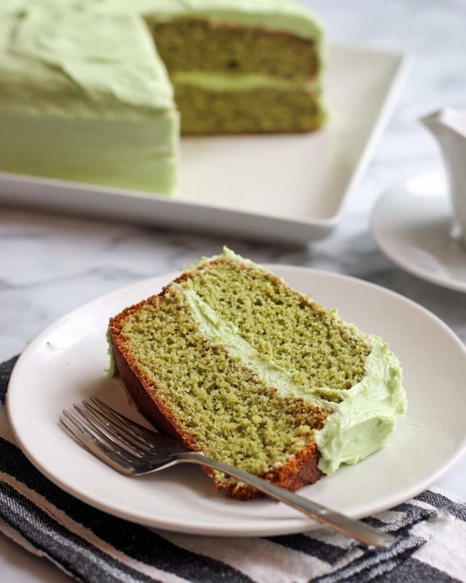 A rectangular loaf cake with a dark brown color sits on a white plate, topped with one thick layer of pale green frosting that has a creamy, swirled texture. The frosting is spread evenly across the top with visible gentle curves and peaks. In front of the plate, a shiny silver cake server is placed on a white marbled surface. Photo taken with an iphone --ar 4:5 --v 7