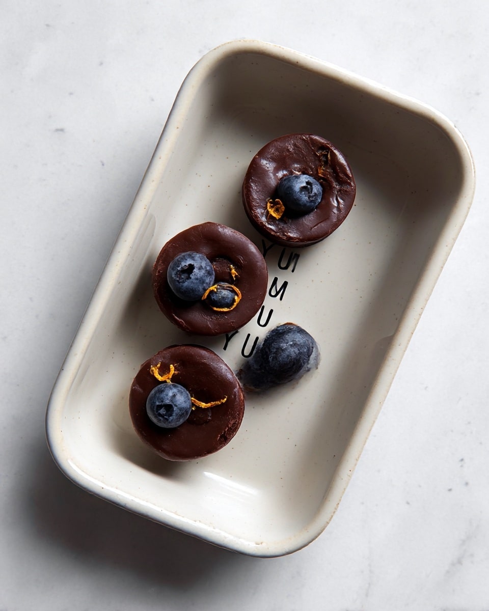 The image shows many small, round chocolate cups filled with whole blueberries and some orange zest bits on top, arranged closely on a white marbled surface. Each chocolate cup has a smooth dark brown color and a slightly shiny texture with blueberries partially embedded, creating lumpy shapes on the surface. Around the chocolate cups, there are loose fresh blueberries scattered randomly, adding a deep blue color contrast. The overall layout is evenly spaced, highlighting the rich, dark color of the chocolate and the vibrant blue of the berries with small hints of orange zest. photo taken with an iphone --ar 4:5 --v 7