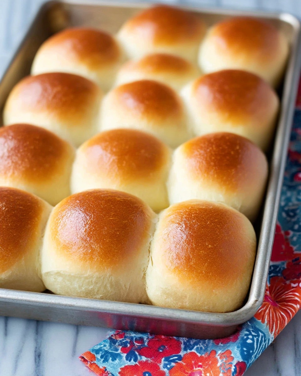 The image shows a metal baking tray filled with twelve soft, round bread rolls arranged in three rows of four. Each roll has a golden brown shiny top with a smooth texture, gradually lightening to a creamy beige color towards the sides. The rolls are touching each other, with slightly puffed, smooth surfaces and a fresh, fluffy look. The tray sits on a white marbled surface, and a colorful cloth with red, blue, and orange floral patterns is partially visible under the tray. Photo taken with an iphone --ar 4:5 --v 7