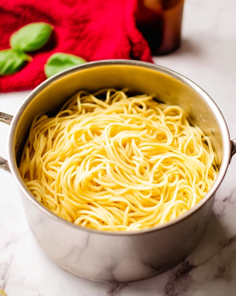 A close-up of a metal pot filled with cooked yellow pasta noodles, neatly piled inside the pot, on a white marbled surface. In the background, there is a red cloth and a brown bottle slightly blurred, with two green basil leaves near the pot. The scene is simple and bright, showing the texture and softness of the pasta strands. Photo taken with an iphone --ar 4:5 --v 7