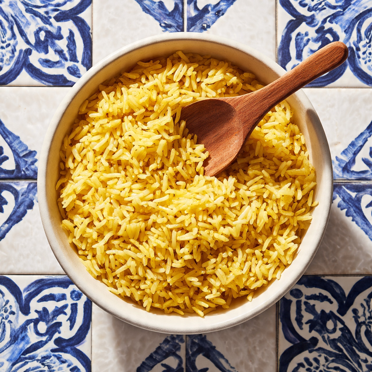 A close-up view of a round white bowl full with yellow cooked rice, each grain visible and fluffy. Inside the bowl, a wooden spoon with a simple brown color lies gently on top, its handle extending upward out of the bowl. The bowl sits on a white marbled surface with blue decorative patterns in a tile-like arrangement in the background. The image is bright and clear, showing the texture of the rice grains well, photo taken with an iphone --ar 4:5 --v 7