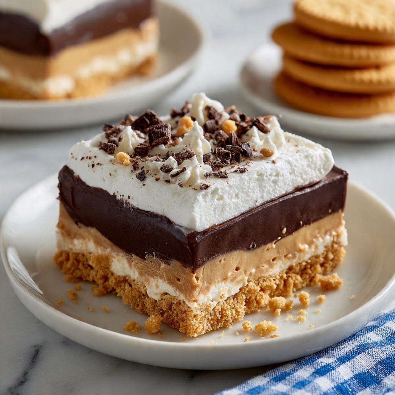 A square piece of layered dessert sits on a white plate on a white marbled surface. The bottom layer is a crumbly, golden brown cookie crust. Above that is a smooth, light brown peanut butter layer, followed by a thick, glossy dark chocolate layer. On top is a fluffy white cream layer sprinkled with small chocolate curls and crushed cookie crumbs. Another similar dessert piece appears blurred in the background on another white plate, with a stack of golden cookies placed nearby. A blue and white checkered cloth is on the right side. photo taken with an iphone --ar 4:5 --v 7