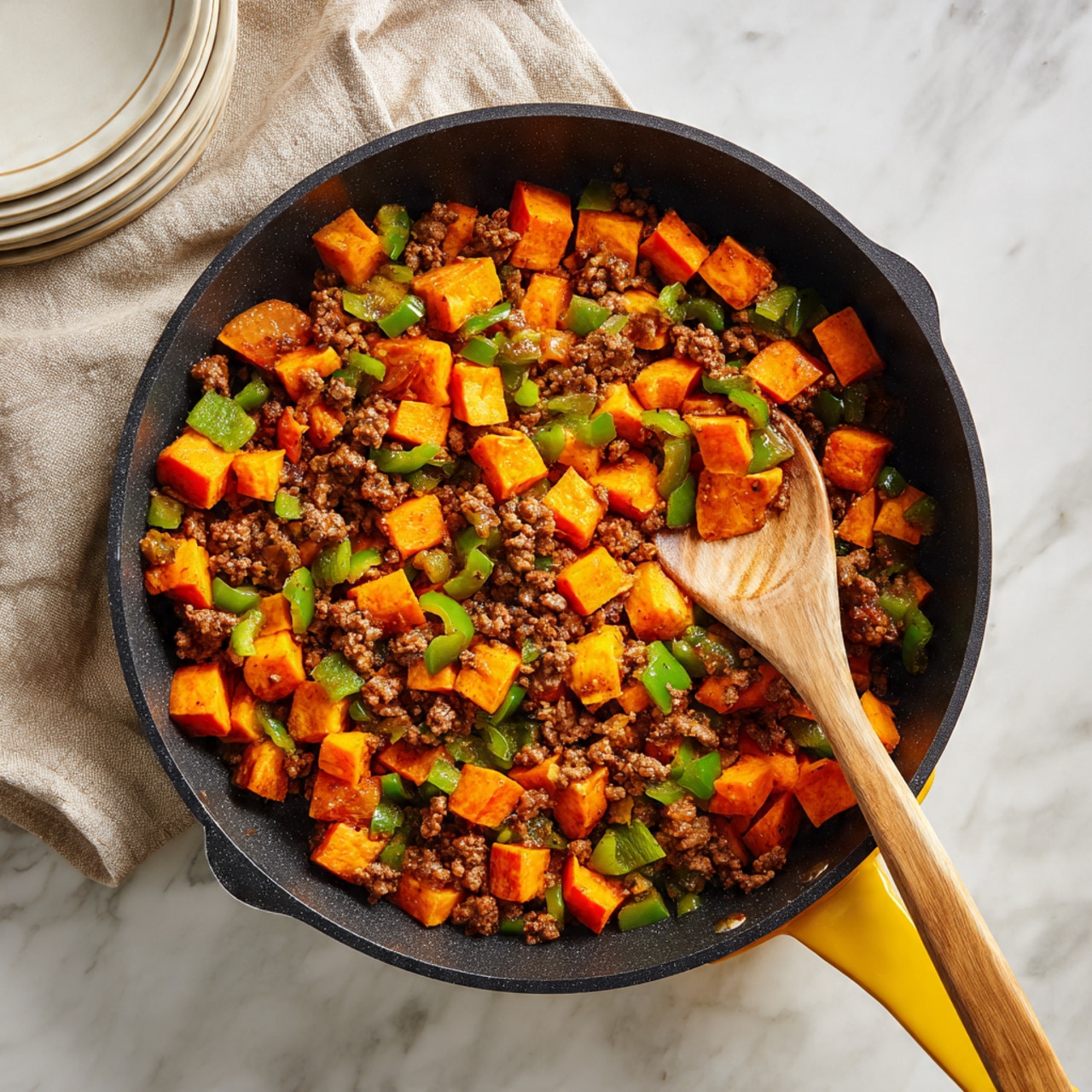 The image shows a black skillet with a yellow handle filled with a cooked dish featuring small cubes of bright orange sweet potatoes, browned ground meat, and chopped green bell peppers mixed together. The sweet potatoes are browned on the edges, giving them a slightly crispy look, and are spread evenly throughout the pan. The green bell peppers add a fresh, crunchy texture contrast with their bright color, scattered evenly as well. A wooden spoon is resting inside the pan, stirring the mixture. The skillet is set on a white marbled surface with a beige cloth nearby, and there is a stack of white plates with a subtle yellow tint on the left side. photo taken with an iphone --ar 4:5 --v 7