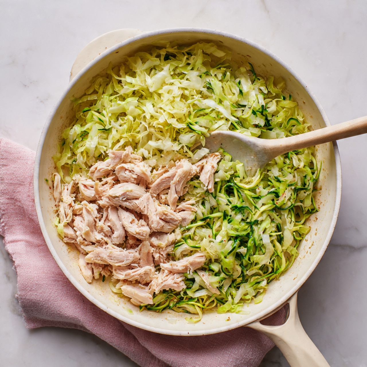A white plate holds a dish with two clear layers. The bottom layer is a bed of small, light brown grains that have a soft, slightly shiny texture, spread evenly across the plate. On top, there is a generous layer of cooked shredded green cabbage mixed with pieces of cooked light tan chicken. The cabbage appears tender and glossy, with thin slices of green and white leaves, and there are small bits of red pepper scattered throughout. The dish is set on a white marbled surface next to a green glass of water, with a beige cloth napkin nearby. photo taken with an iphone --ar 4:5 --v 7