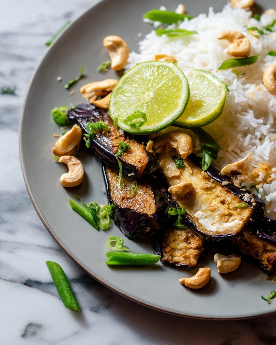 A white plate holds two main layers: on the left, a pile of soft white rice with a fluffy texture, and on the right, golden brown roasted eggplant pieces with dark, slightly crispy edges. On top of both layers are scattered creamy-colored cashew nuts and thinly sliced bright green spring onions. Two lime wedges, light green with a glossy texture, sit on top of the rice near the roasted eggplant. In the background, there is a small metal cup filled with more sliced green onions, all placed on a white marbled surface. photo taken with an iphone --ar 4:5 --v 7