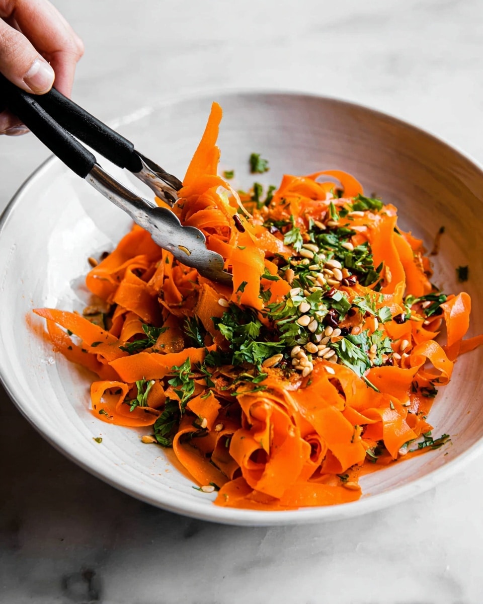 A white shallow bowl holds a bright orange dish made of thin, wide carrot ribbons that resemble noodles. The carrot ribbons are mixed and piled loosely, showing a slightly shiny texture from light dressing or oil. On top, fresh chopped green herbs and small brown seeds are scattered, adding contrast and texture. A woman's hand uses black metal tongs to lift some carrot ribbons from the bowl. The bowl sits on a white marbled surface. photo taken with an iphone --ar 4:5 --v 7