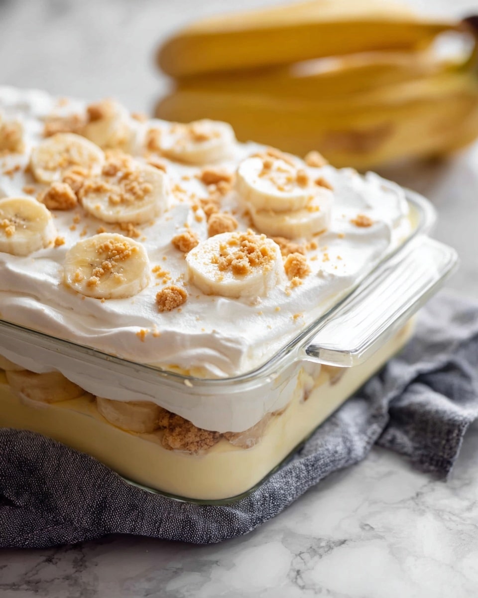 This image shows a glass rectangular dish filled with a layered banana dessert placed on a white marbled surface, partly resting on a gray cloth. The bottom layer appears creamy and pale yellow in color, topped by slices of banana arranged in a single layer along the edges. Above the bananas, there is another creamy layer, lighter in color, followed by a thick white whipped topping that covers the surface. The top is decorated with scattered banana slices and crushed light brown cookie pieces, adding texture and color contrast. In the background, slightly out of focus, a bunch of ripe bananas is visible. Photo taken with an iphone --ar 4:5 --v 7