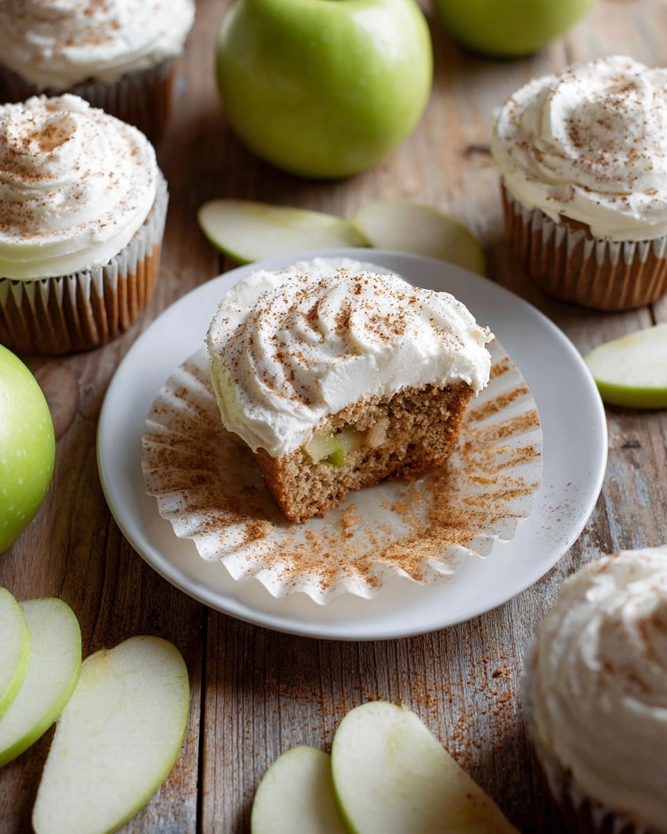 The image shows a rusty metal baking tray holding eleven round cupcakes. Each cupcake has two layers: the base layer is a soft, golden-brown cake, and the top layer is a thick swirl of white creamy frosting, sprinkled with a light dusting of brown cinnamon powder. The tray is placed on a wooden texture background with a green apple in the top left corner and a sliced green apple on the bottom right. photo taken with an iphone --ar 4:5 --v 7