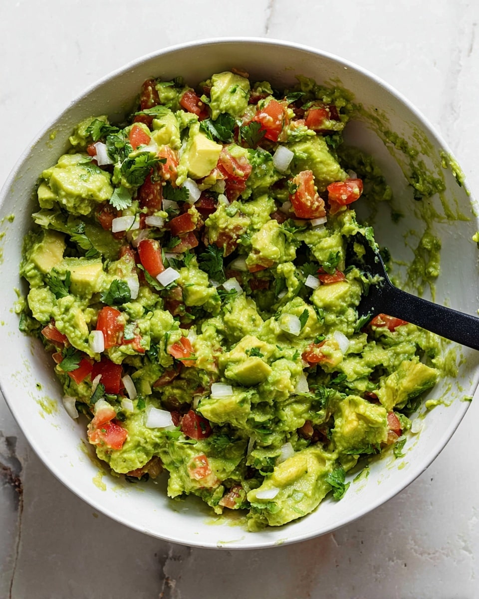 A white bowl holds a fresh mixture of guacamole showing three main layers: bright green mashed avocado chunks with a soft, creamy texture forms the base, scattered with small, bright red tomato pieces adding a firm, juicy contrast, and finely chopped white onions mixed evenly, providing a crisp texture. Small bits of dark green cilantro leaves are spread throughout, adding a fresh look. A black spoon is placed inside the bowl on the right side, resting on the mixture. The bowl is set on a surface with a white marbled texture photo taken with an iphone --ar 4:5 --v 7
