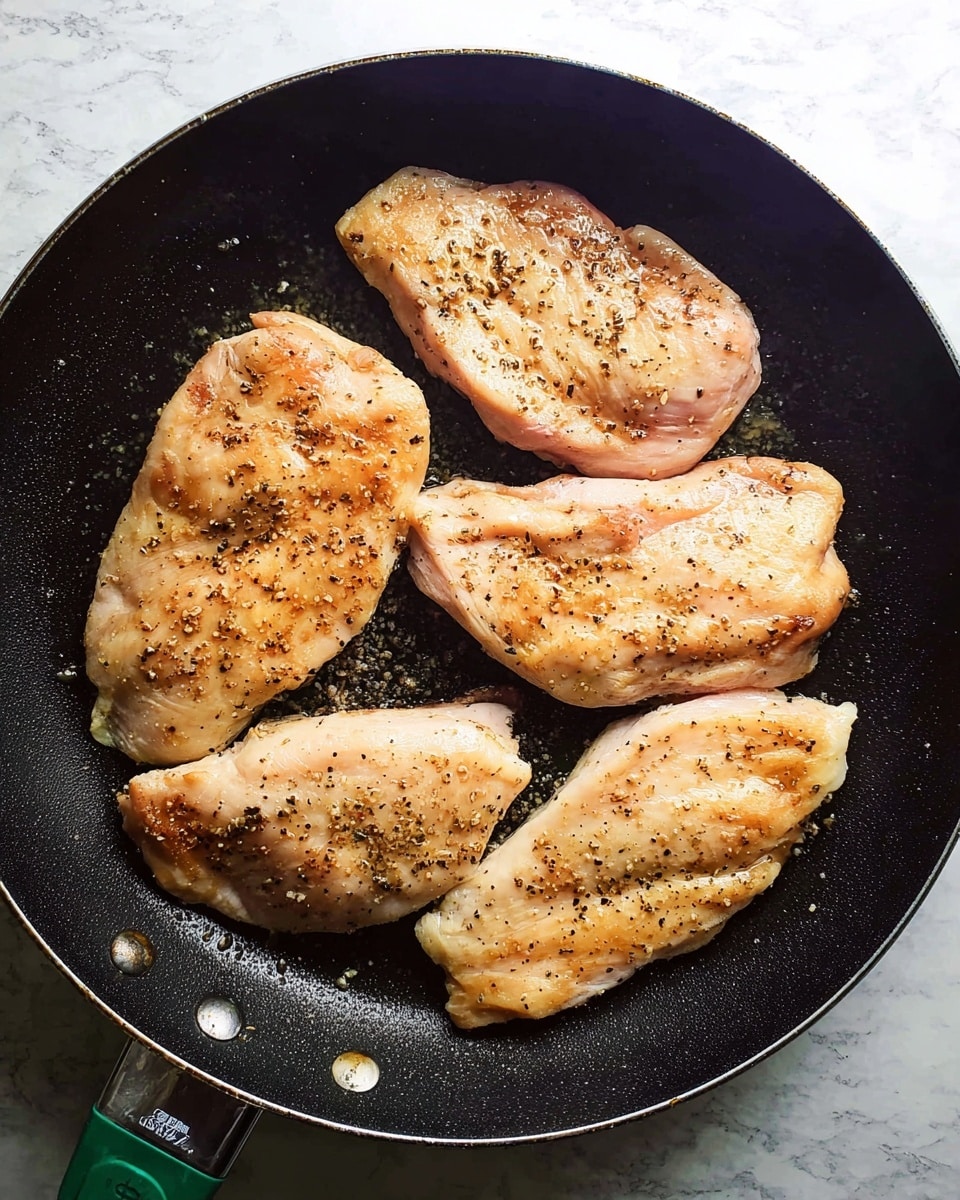 A dark black pan with a green handle holds four pieces of light pink chicken placed flat in the pan in a close layout. Each piece of chicken is sprinkled with coarse brown seasoning and black pepper, showing a slightly shiny texture with a little bit of oil on the surface. The background consists of a white marbled texture. photo taken with an iphone --ar 4:5 --v 7