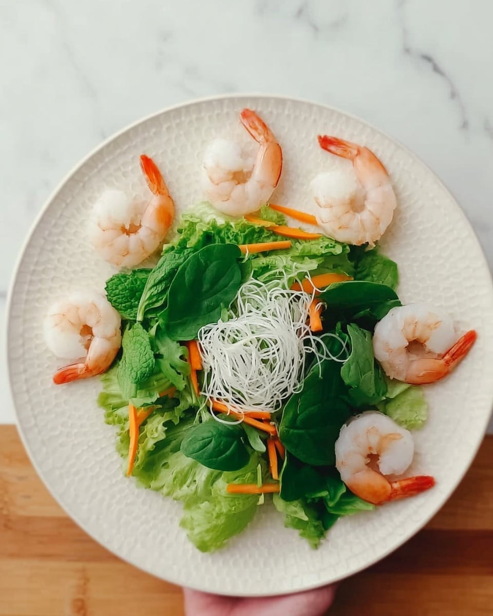 A white round plate is arranged with five peeled shrimp placed in a semi-circle along the upper edge of the plate, showing their light pink and white soft texture. Below the shrimp, in the center of the plate, there is a bed of mixed fresh green leaves with smooth and curly edges, topped with thin white noodles that have a smooth and slightly shiny texture. Bright orange carrot strips are woven through the noodles, adding a splash of color. The plate is on a white marbled surface. A woman's hand is holding the plate from the right edge. photo taken with an iphone --ar 4:5 --v 7