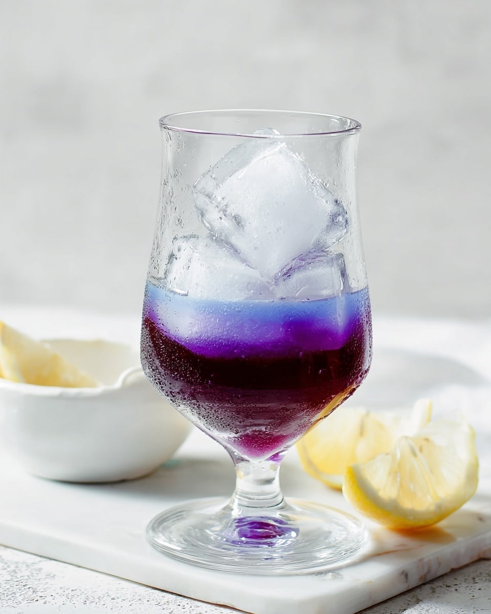A clear glass holds a drink with two layers: a thick deep purple liquid at the bottom and three large clear ice cubes stacked above it. The glass has a rounded base that narrows near the top, showing droplets of condensation. It stands on a white marbled surface with a white small bowl and two lemon wedges nearby. The background is also white and softly lit. photo taken with an iphone --ar 4:5 --v 7