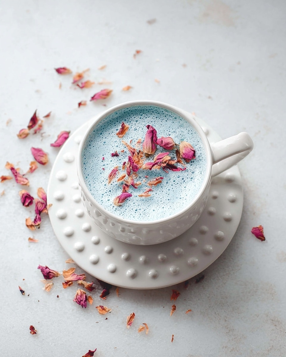 A frothy light blue drink fills a vintage white cup with rounded bumps around the rim and handle, topped with dried pink and yellow rose petals scattered mostly on one side of the foam. The cup sits on a matching white saucer with similar rounded bumps along the edge, all placed on a white marbled surface. Nearby, there is a white bowl filled with deep blue powder and scattered dried petals adding soft touches of pink and beige around. Photo taken with an iphone --ar 4:5 --v 7