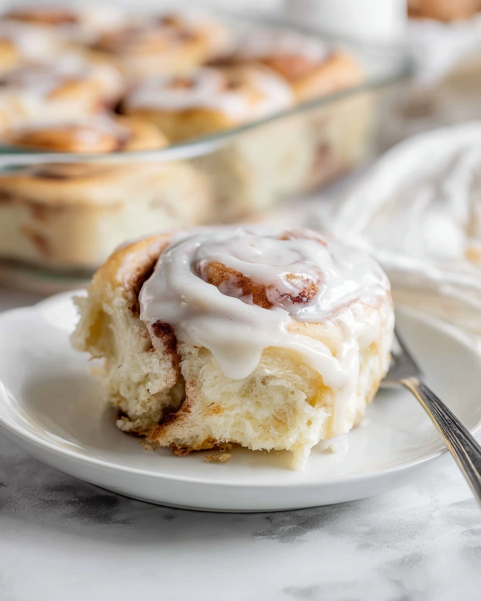 The image shows a close-up of one cinnamon roll on a white plate on a white marbled surface. The cinnamon roll has three main layers: a soft, fluffy dough base with a light golden color, a middle layer with a swirl of cinnamon brown, and a top layer thickly covered with smooth, white icing that slightly drips down the sides. In the background, more cinnamon rolls sit in a clear glass baking dish, also topped with white icing. The scene has soft natural light, making the textures of the dough and icing clear and inviting photo taken with an iphone --ar 4:5 --v 7