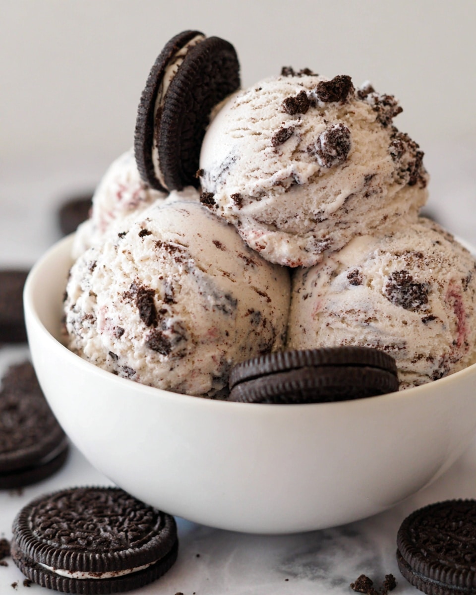 A close-up view of a single scoop of cookies and cream ice cream served in a white bowl with a textured exterior. The ice cream is pale cream in color with small black crumbs and occasional small pink specks mixed throughout. Two whole chocolate sandwich cookies with cream filling are placed upright behind the ice cream scoop, partially submerged. The bowl sits on a white marbled surface, and part of another cookie is visible at the bottom edge of the image. photo taken with an iphone --ar 4:5 --v 7