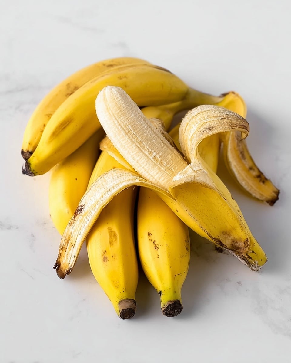 A close-up image shows a woman's hand holding a small rolled crepe filled with soft, mashed banana that has a creamy yellow color and smooth texture with some small brown spots. In the background, two more rolled crepes sit on a white plate with thin, dark chocolate syrup lines artistically drizzled in a criss-cross pattern. The plate is placed on a white marbled surface. Behind, there are a few ripe bananas with brown spots and a glass jar with red roses and green leaves, slightly out of focus. The soft light highlights the textures and colors. photo taken with an iphone --ar 4:5 --v 7