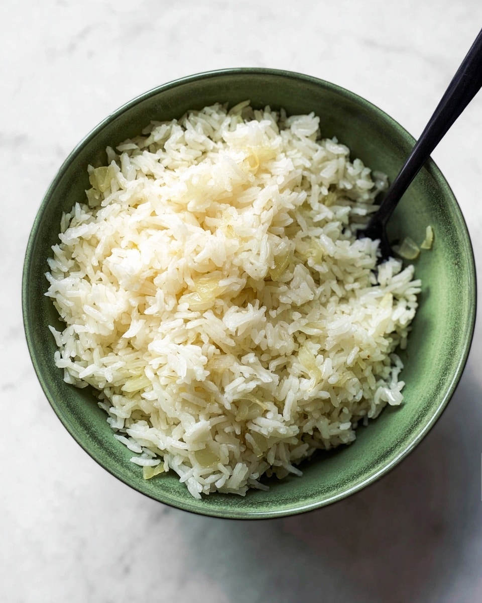 A single green bowl filled with cooked white rice grains that are fluffy and slightly shiny, showing some small soft pieces of cooked onion mixed evenly throughout. A black spoon is placed inside the bowl on the right side, partially buried in the rice. The bowl sits on a white marbled surface. photo taken with an iphone --ar 4:5 --v 7