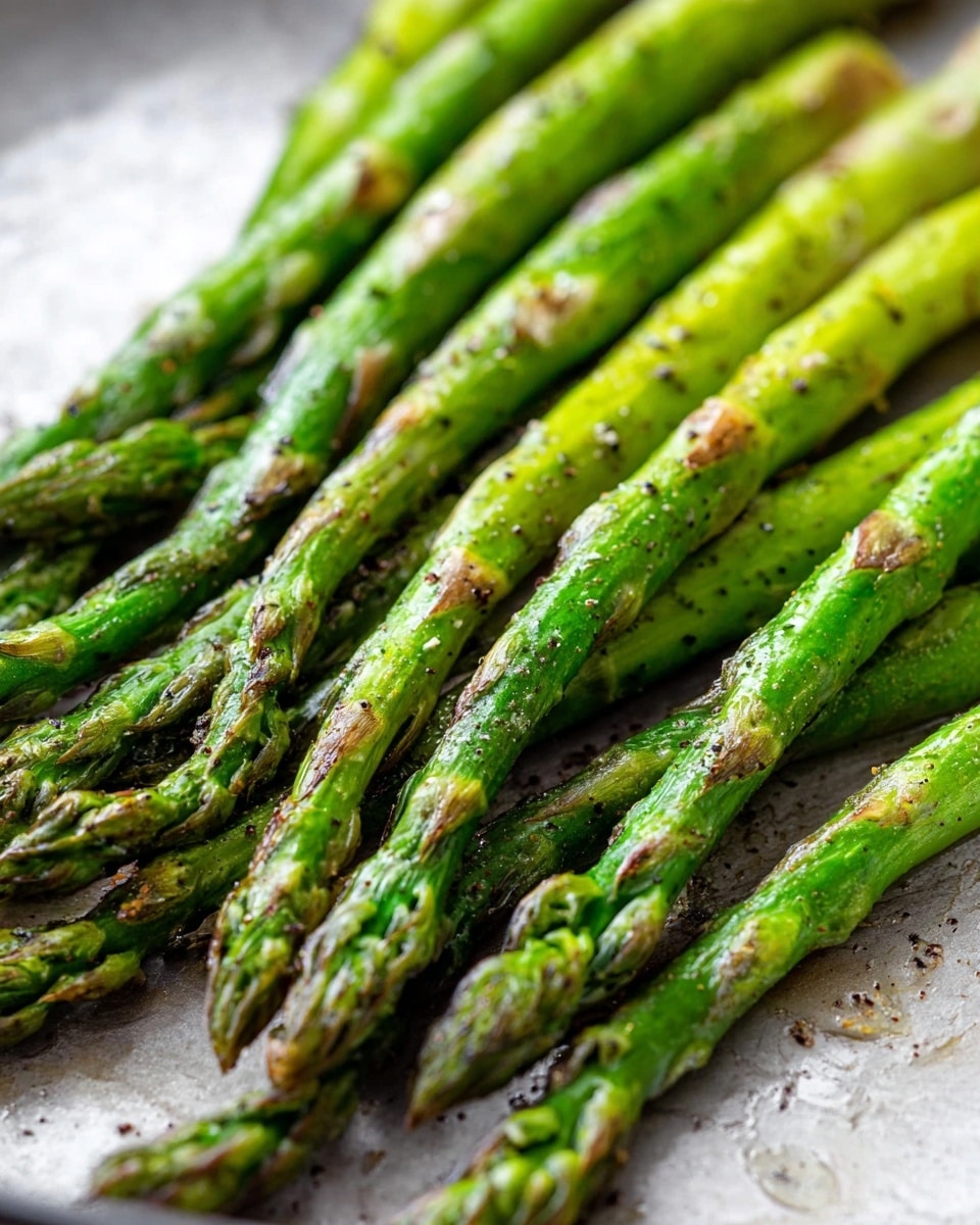 The image shows bright green asparagus spears cooked with some light brown roasting marks, placed close together on a pan with a light, slightly textured surface. The asparagus looks slightly shiny from oil and has small specks of black pepper on it. The spears have a fresh and tender appearance with visible texture from cooking, some tips are more browned than others. The background is a white marbled texture. Photo taken with an iphone --ar 4:5 --v 7