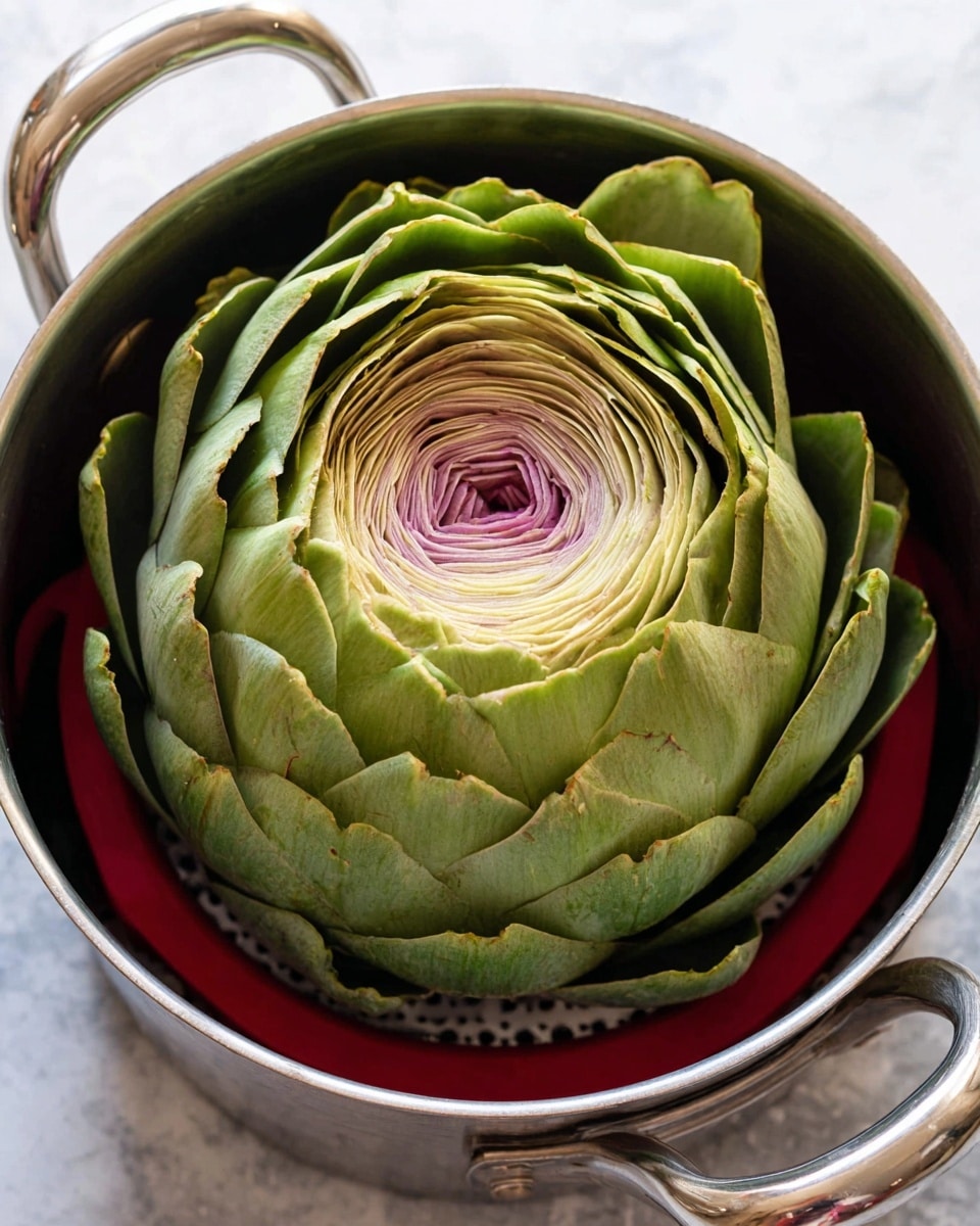 A large artichoke sits in the center of a silver metal pot with two handles. The artichoke has many layers of tightly packed leaves. The outer leaves are a bright green, and as you look toward the middle, the leaves become lighter green, then pale yellow, and finally a soft purple-gray at the core. The artichoke rests on a red steamer basket inside the pot. The surface below the pot is white marbled texture photo taken with an iphone --ar 4:5 --v 7