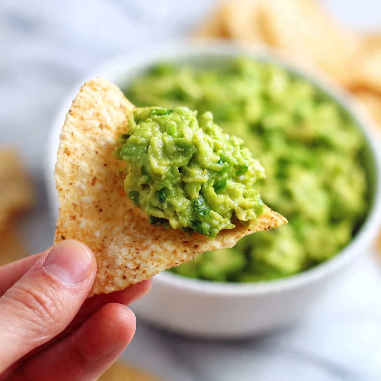 The image shows a white bowl filled with chunky green guacamole topped with sliced green onions. The guacamole has a varied texture with visible small lumps and bits of avocado and herbs. The bowl sits on a white marbled surface with tortilla chips placed nearby on the right side, and a halved lime and a small sprig of dried lavender on the left. The background includes a white container and some white ramekins on a wooden board, softly blurred to keep focus on the guacamole. photo taken with an iphone --ar 4:5 --v 7