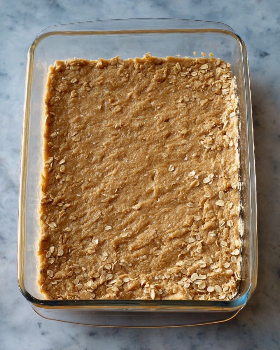 Six square snack bars are shown on a dark surface with a white marbled texture. Each bar has two layers: the bottom layer is a crumbly, rough-textured golden brown base, and the top layer is a smooth, creamy pale beige frosting spread evenly but with gentle swirls. The closest bar in the foreground has a small bite taken out of its corner, revealing the soft inside of the base. Small crumbs are scattered around the bars, enhancing the textured look. The photo taken with an iphone --ar 4:5 --v 7
