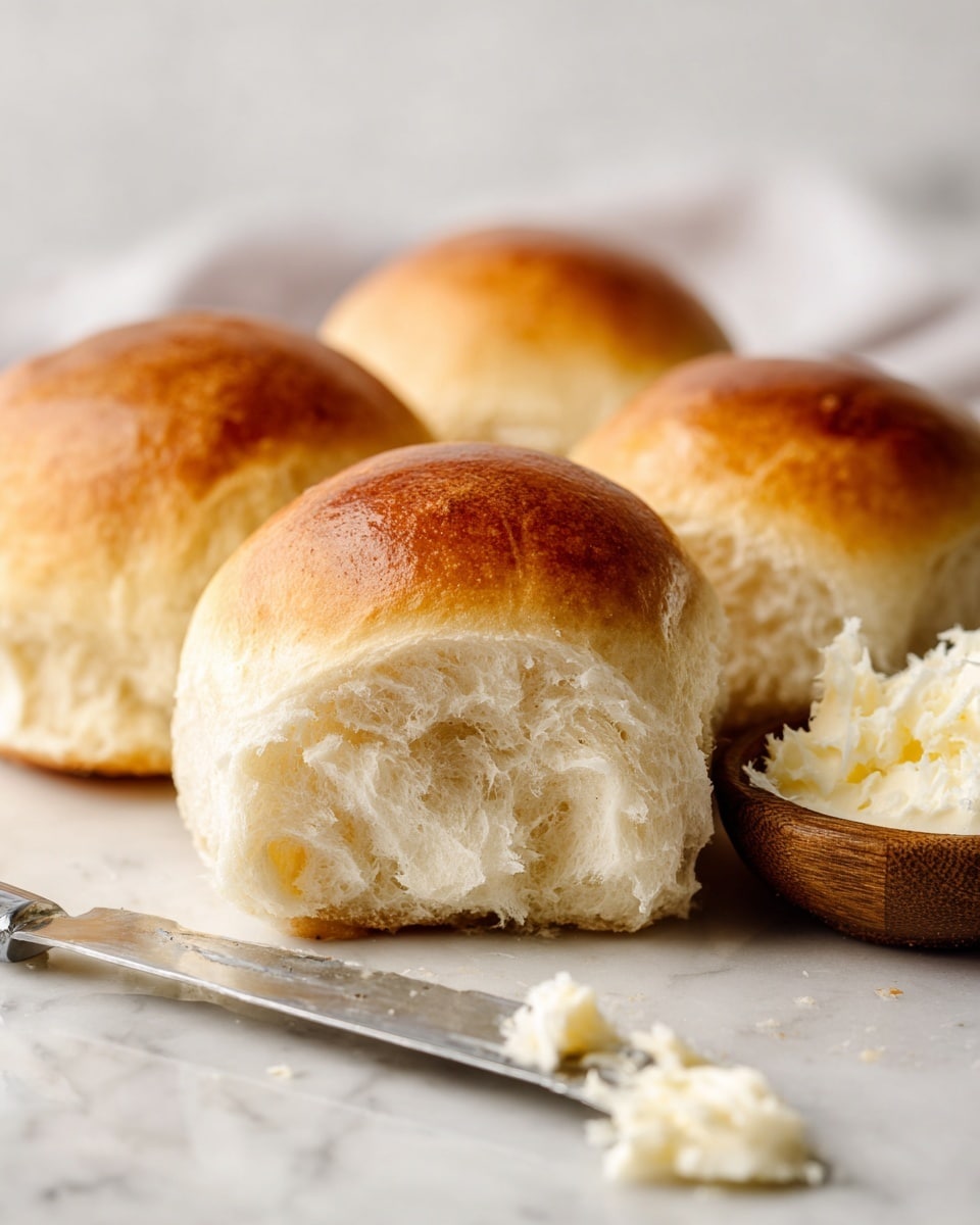 The image shows four soft dinner rolls with a golden brown top and a light, fluffy inside. They are close together on a white marbled surface, with one roll prominently in the front center showing its airy texture. To the right, there is a small wooden bowl filled with white creamy butter or spread. In front of the rolls and bowl, a silver butter knife lies flat on the white marbled surface with a small smear of butter near its tip. The background is softly blurred, highlighting the rolls. photo taken with an iphone --ar 4:5 --v 7