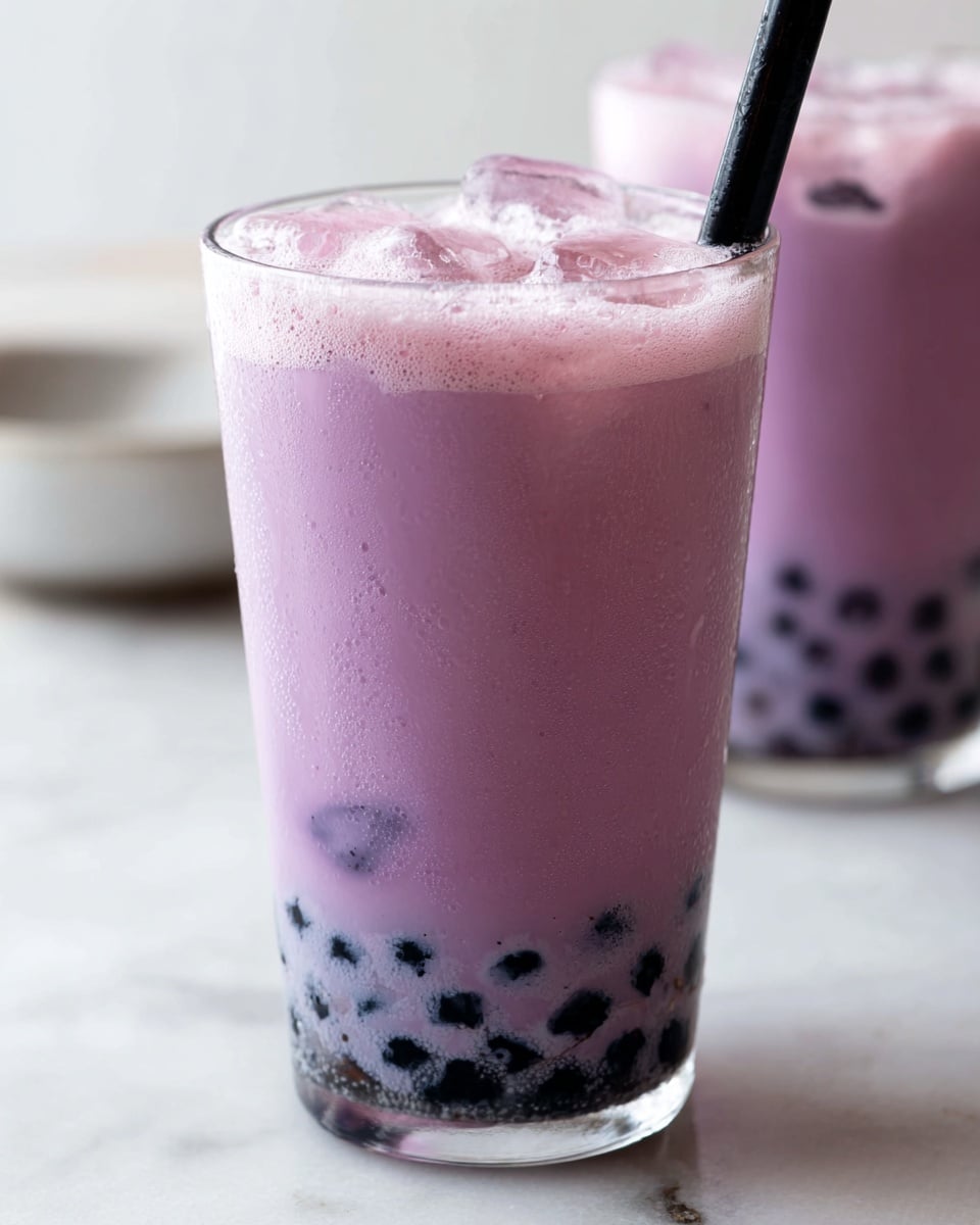 A clear glass filled with three layers: at the bottom, dark black round tapioca pearls; the middle layer is a smooth, light purple milk tea with visible ice cubes inside; the top layer shows a frothy, pale pink foam with small bubbles. Two black straws stick out from the glass. The glass sits on a white marbled surface with a blurred second glass of the same drink in the background. photo taken with an iphone --ar 4:5 --v 7