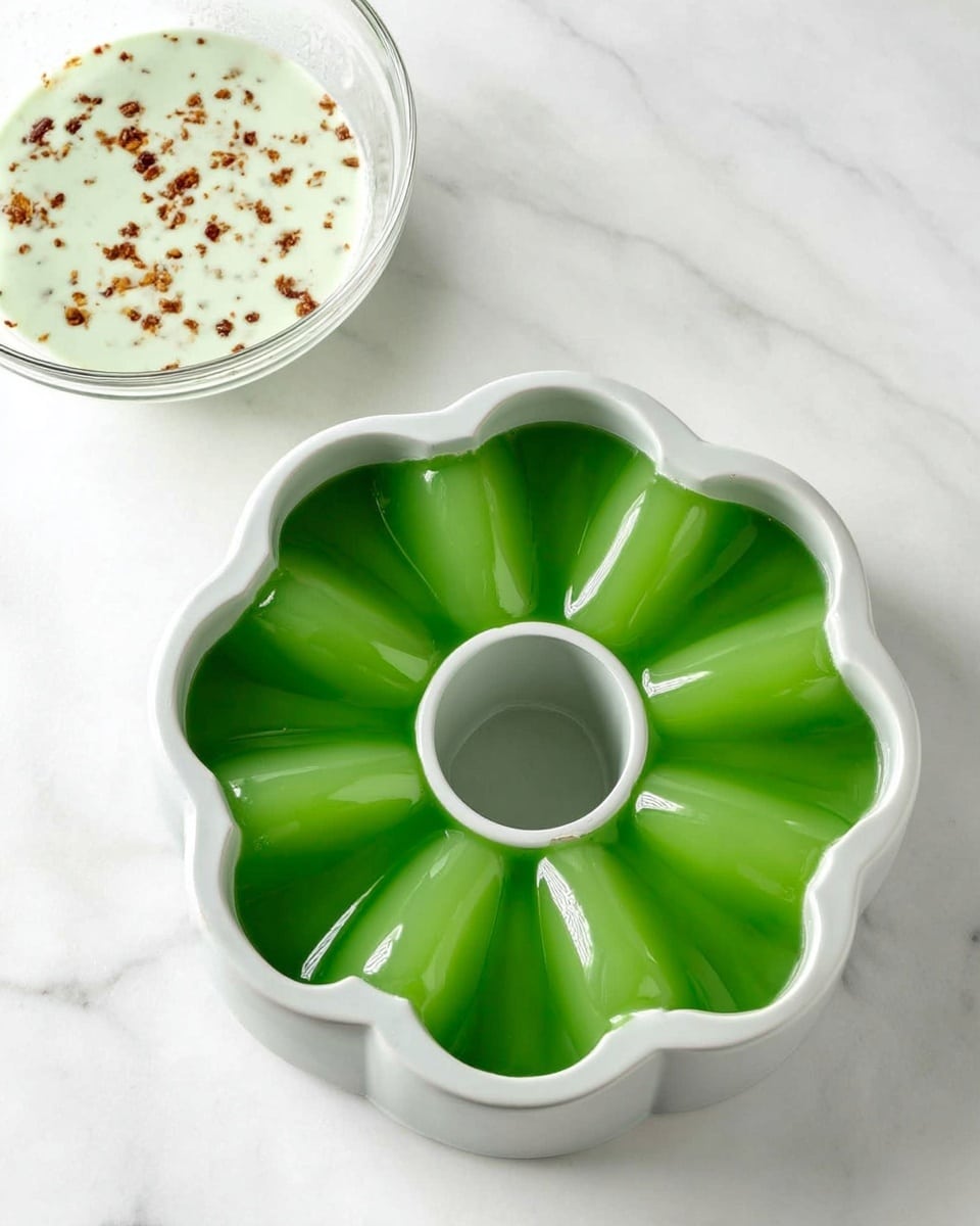 The image shows a green gelatin dessert shaped like a ring with a smooth, shiny top layer. Inside the ring is a thick, light green creamy layer mixed with small brown pieces, giving it a textured look. A slice has been taken out and placed on a white plate with a wavy edge, showing the two layers clearly. The dessert sits on a white marbled surface with a few whole limes scattered nearby, and there is a blurred white bowl filled with limes in the background. Photo taken with an iphone --ar 4:5 --v 7