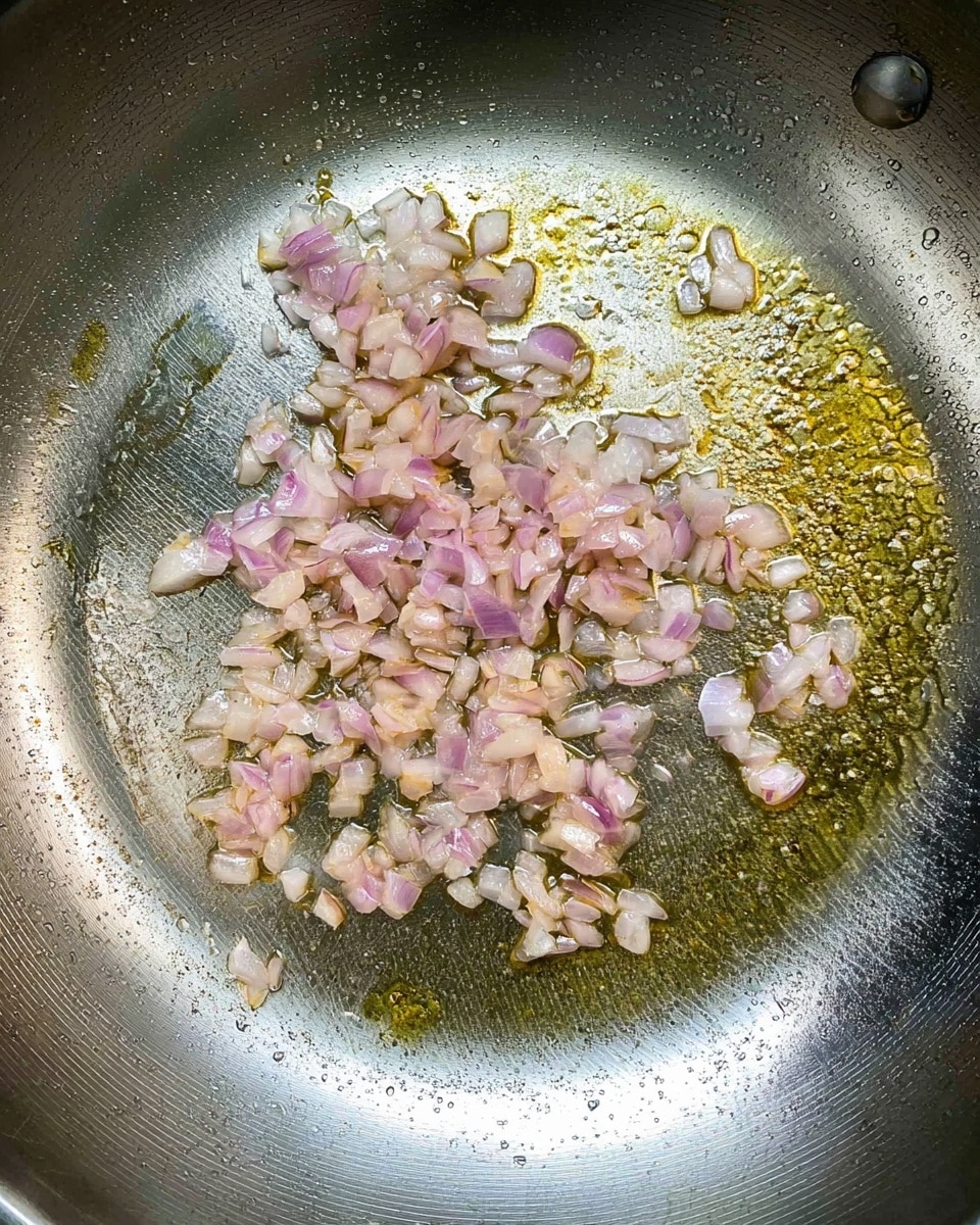 A shiny silver pan is shown from above with small pieces of chopped light pink shallots cooking in clear golden oil. The shallots are spread out mostly in the upper left part of the pan, slightly translucent with some browning edges. The pan surface has a metallic texture with some oil glistening and tiny bubbles around the shallots. photo taken with an iphone --ar 4:5 --v 7
