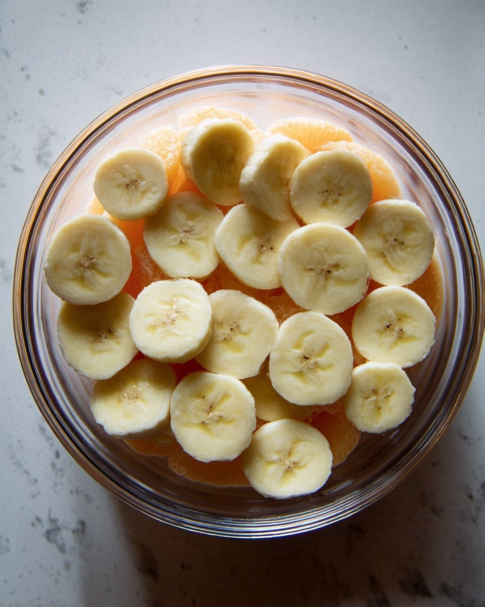 The image shows a clear glass bowl filled with two layers. The bottom layer is made of round, light orange slices, while the top layer is made of round, pale yellow banana slices. The bowl is placed on a white marbled surface, and the top view captures the details clearly, showing the smooth texture of both fruit layers, with the banana slices slightly overlapping each other. photo taken with an iphone --ar 4:5 --v 7