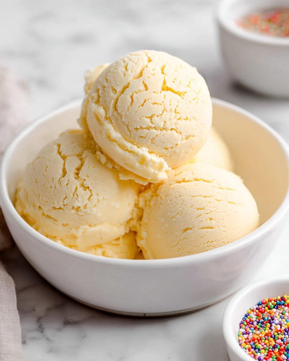 Three scoops of light creamy yellow ice cream with a smooth yet slightly cracked texture are placed closely together in a white bowl. The top scoop is centered and slightly raised above the other two, showing fine lines on its surface. The bowl sits on a white marbled surface with a small white bowl filled with colorful round sprinkles placed partially visible in the lower right corner. Photo taken with an iphone --ar 4:5 --v 7