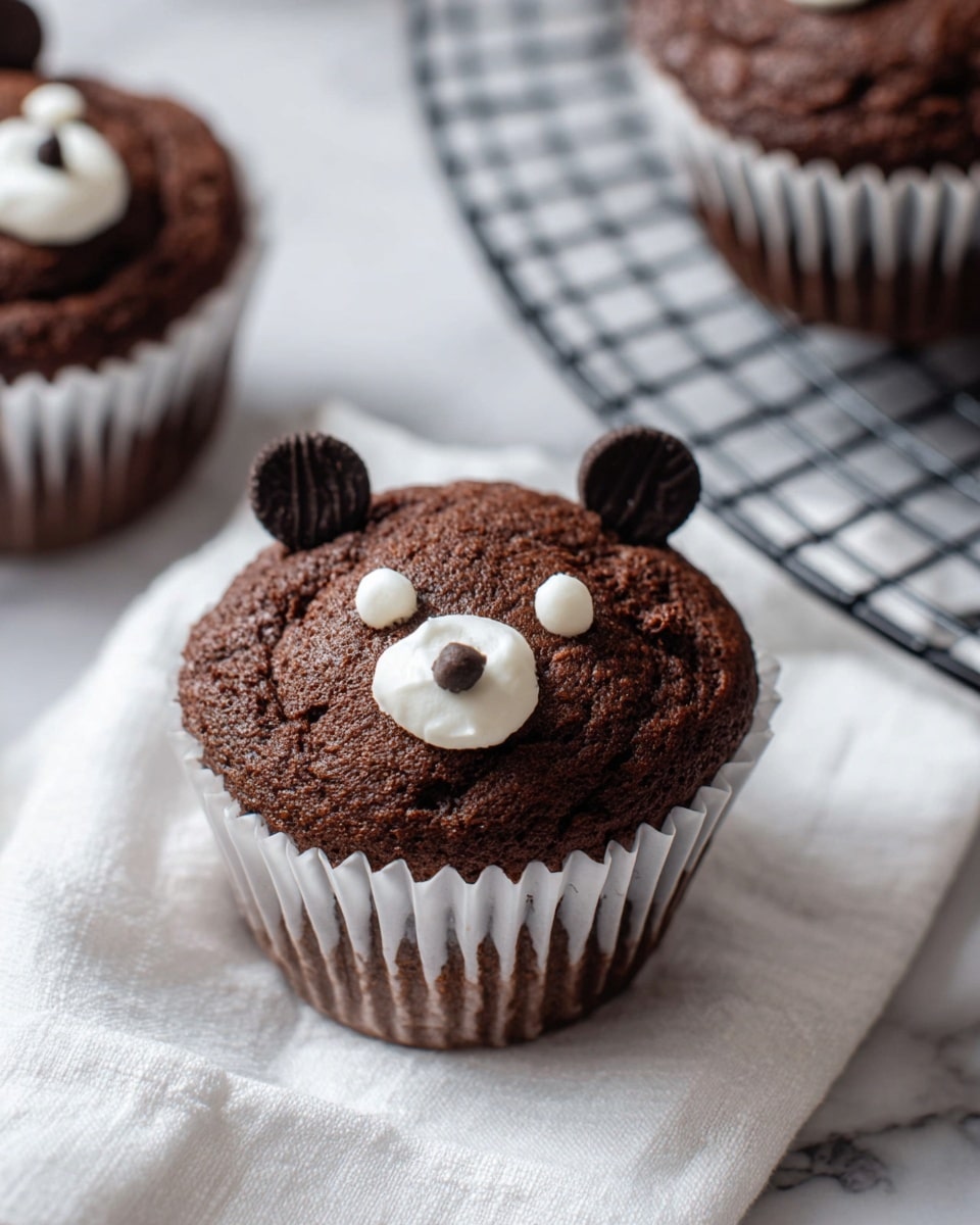 This image shows several chocolate cupcakes with a soft, cracked brown top placed on a round metal cooling rack. Each cupcake is lined with white paper. On the top of every cupcake, there are small decorations made of white and dark chocolate pieces that look like a bear's face, with two dark chocolate circles on top for ears and a white dollop in the center resembling a nose, plus tiny black dots for eyes on the white marbled surface. The background is also white marbled texture and a white cloth is softly draped nearby. photo taken with an iphone --ar 4:5 --v 7