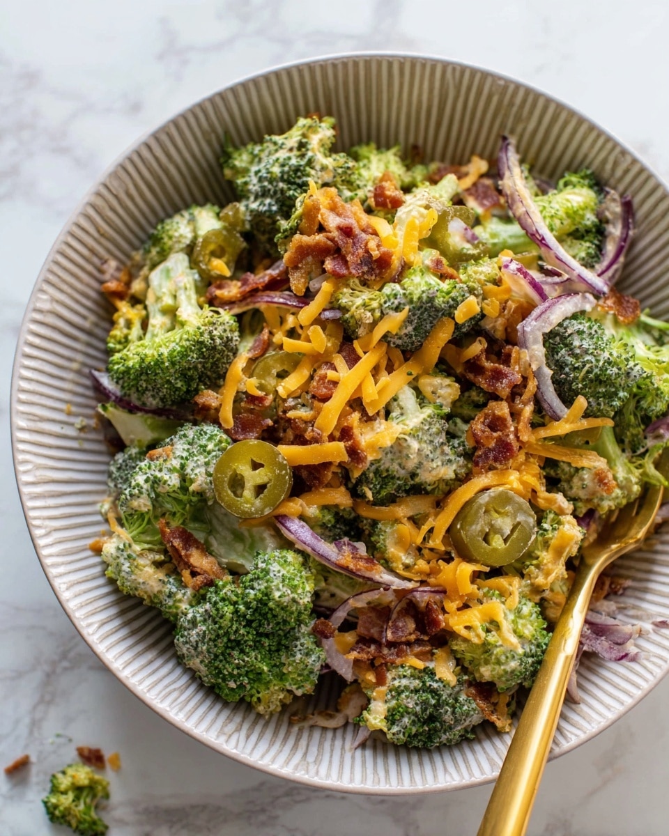 A white bowl filled with a layered broccoli salad on a white marbled surface; the first layer is dark green broccoli florets mixed with light green broccoli stems all mixed in a creamy white dressing; scattered over this are small pieces of reddish-brown bacon bits and bits of chopped red onion; on top there are bright orange shredded cheddar cheese pieces and round slices of yellow-green pickled jalapeños creating vibrant spots of color; a silver spoon with a long handle sits inside the bowl along one edge; photo taken with an iphone --ar 4:5 --v 7