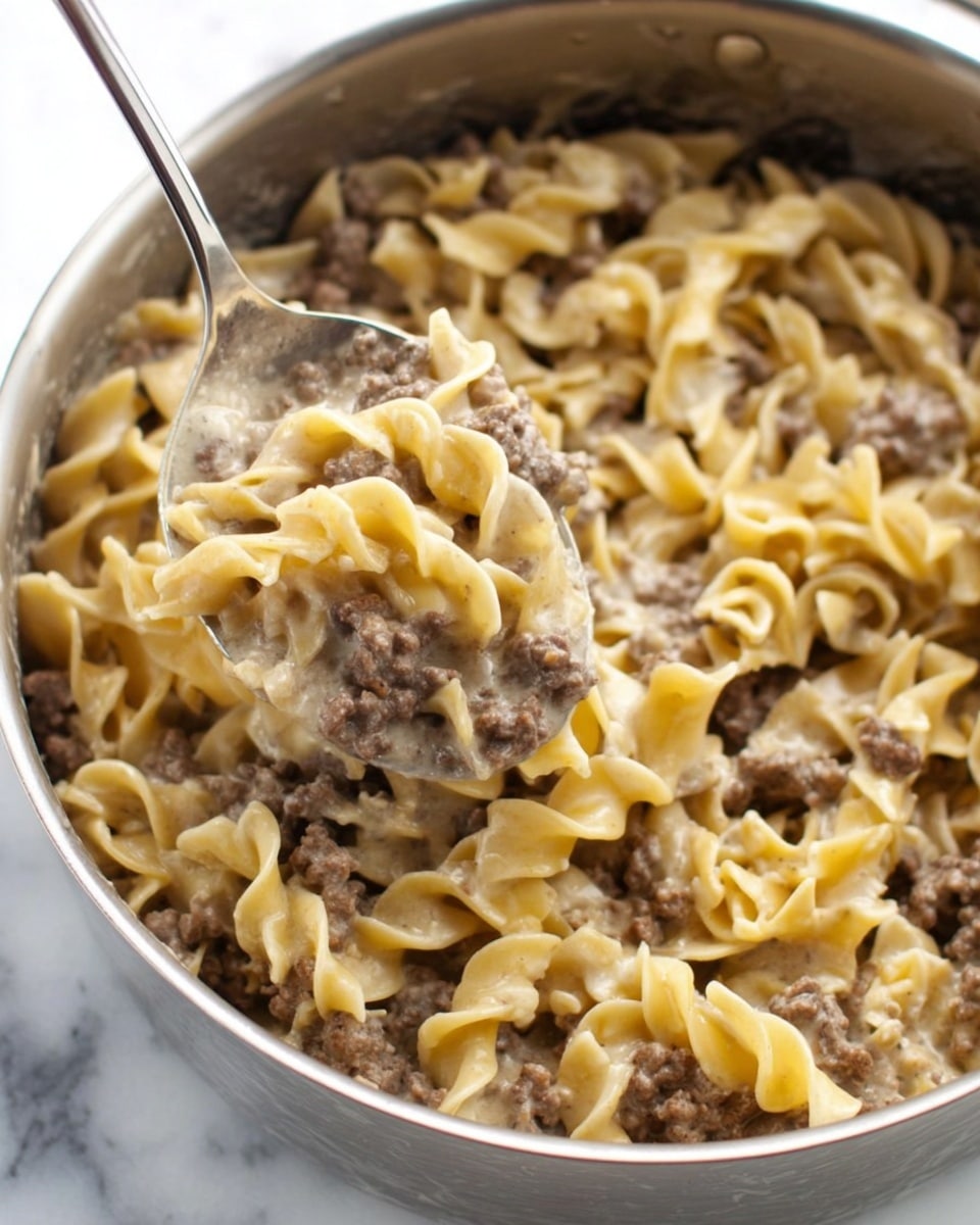 The image shows a close-up view of a large silver pan filled with a dish made of two main layers. The bottom layer consists of cooked curly egg noodles showing a yellowish color with hints of light seasoning. The top layer is cooked ground beef, mixed evenly with the noodles, appearing brown and slightly crumbly. The textures of the noodles are soft and wavy, contrasting with the rough, meaty texture of the beef. The pan sits on a white marbled surface. Photo taken with an iphone --ar 4:5 --v 7