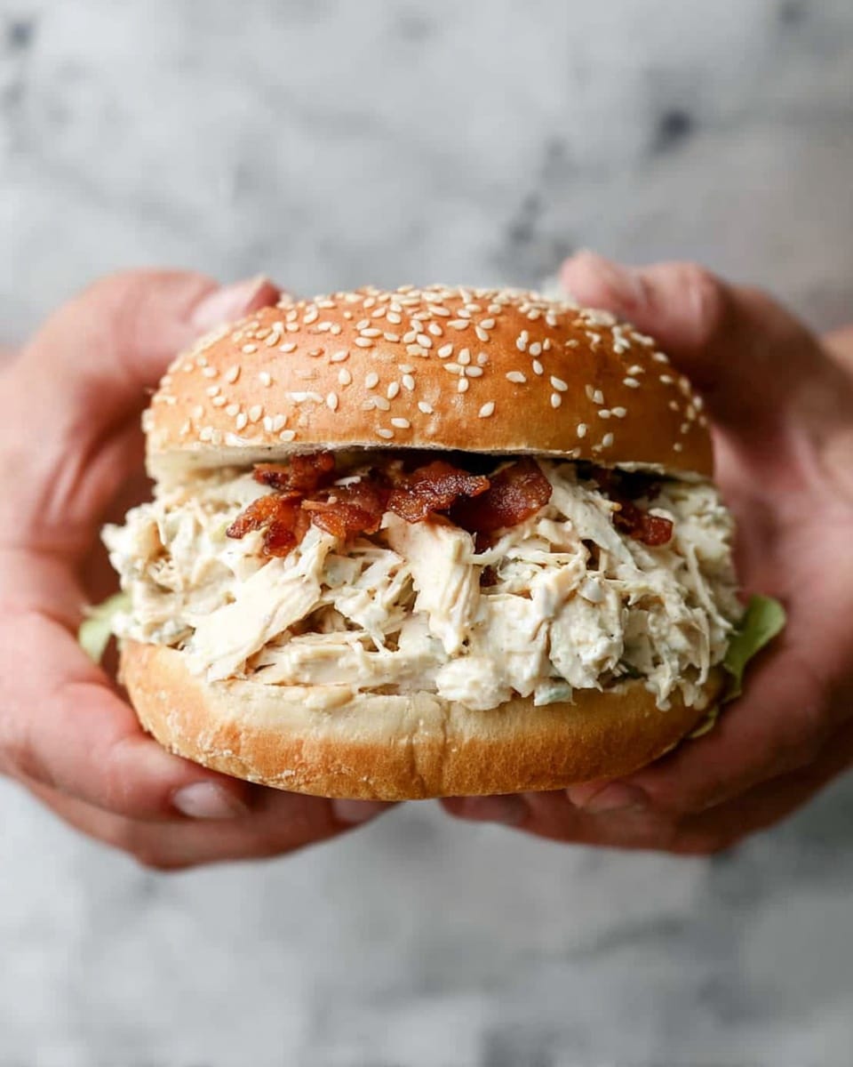 A sandwich inside a white sesame seed bun with two layers: the bottom layer is creamy shredded chicken salad mixed with herbs and mayonnaise, light beige in color with a smooth texture, the middle layer has crispy cooked bacon pieces in dark reddish-brown, scattered on top of the chicken salad, and the top sesame seed bun covers the sandwich. The sandwich rests in a blue basket lined with white parchment paper on a white marbled surface. A white plastic knife is visible behind the sandwich. Photo taken with an iphone --ar 4:5 --v 7