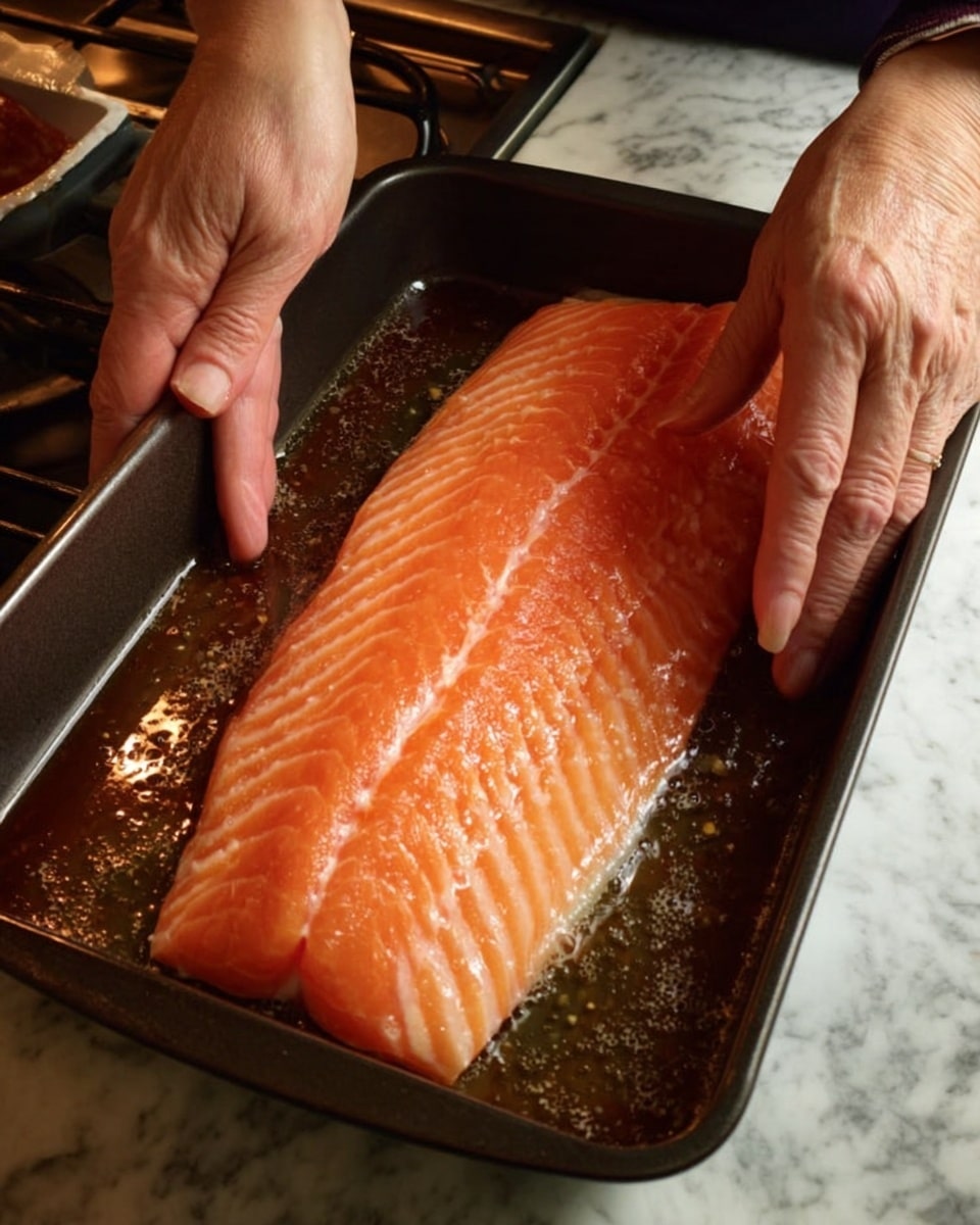 A large, fresh salmon fillet with a smooth, shiny orange skin and light white stripes is placed in a dark roasting pan filled with a dark liquid marinade, with two woman's hands gently pressing its edges. The pan surface is slightly worn and the background shows a kitchen counter with appliances, all resting on a white marbled texture. photo taken with an iphone --ar 4:5 --v 7