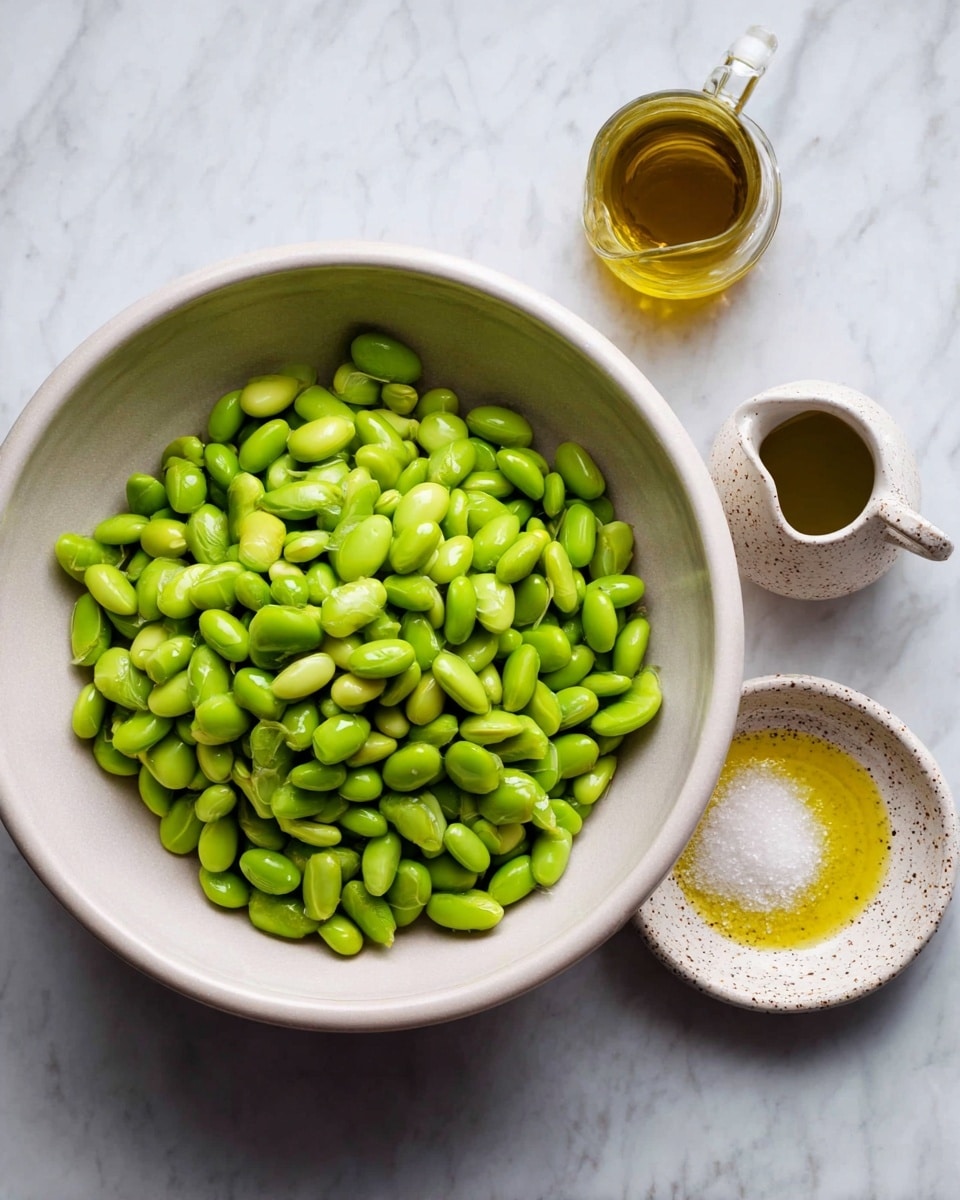 A large white bowl filled with bright green shelled edamame beans sits on a white marbled surface. To the right of the bowl, there are two small white speckled dishes—one round dish holds a small pile of white salt, and a small pitcher contains golden olive oil. The scene is simple and clean, showing fresh ingredients ready for preparation. Photo taken with an iphone --ar 4:5 --v 7