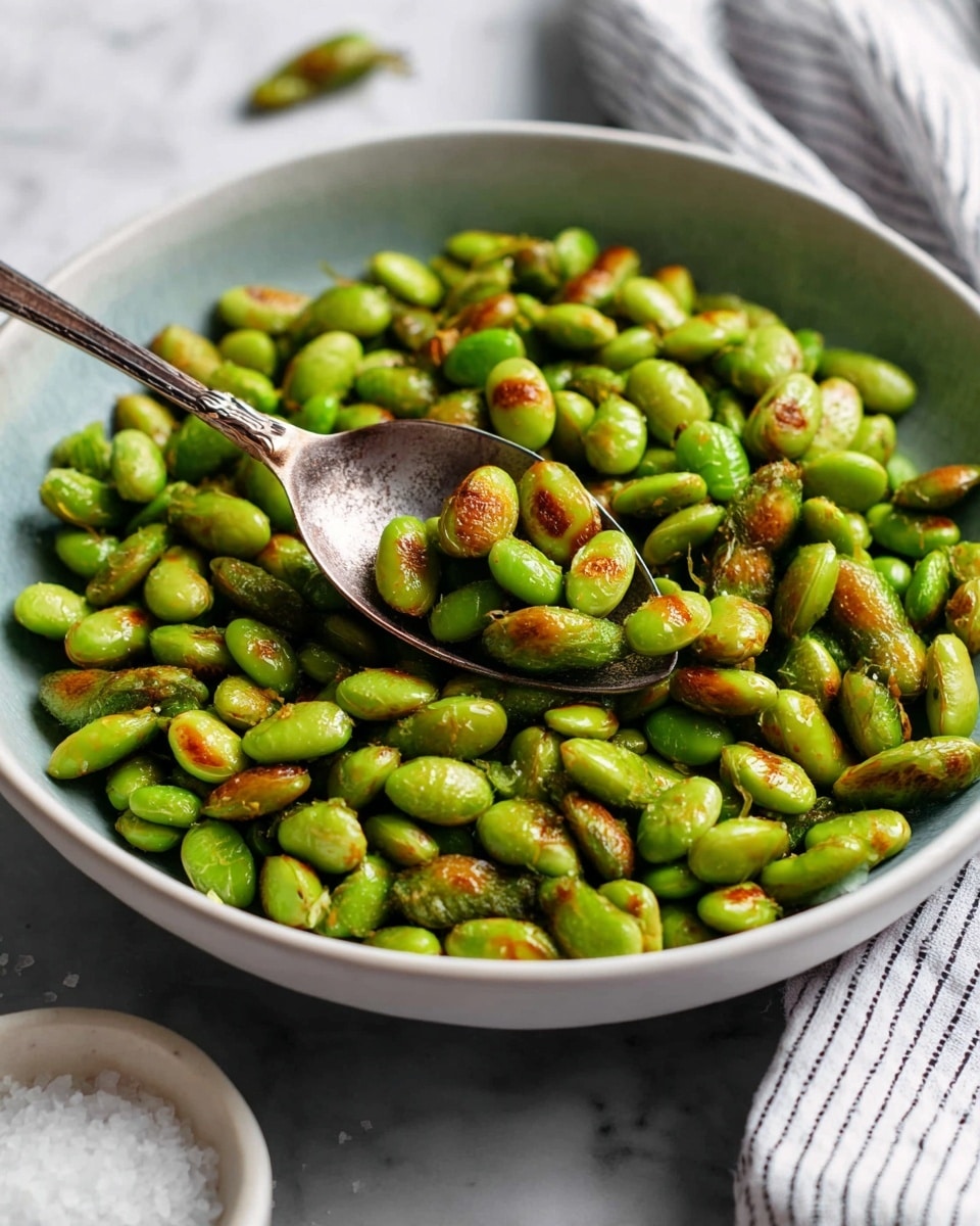 A close-up view of a clear glass jar filled with bright green roasted edamame beans, some with slight golden brown spots, showing a slightly shiny and textured surface. A few edamame beans are scattered near the jar on a white marbled surface. In the background, there is a round white plate with a green and yellowish edge, slightly blurred. The jar has a metal clasp on the side, and the whole scene is well-lit with natural light. photo taken with an iphone --ar 4:5 --v 7
