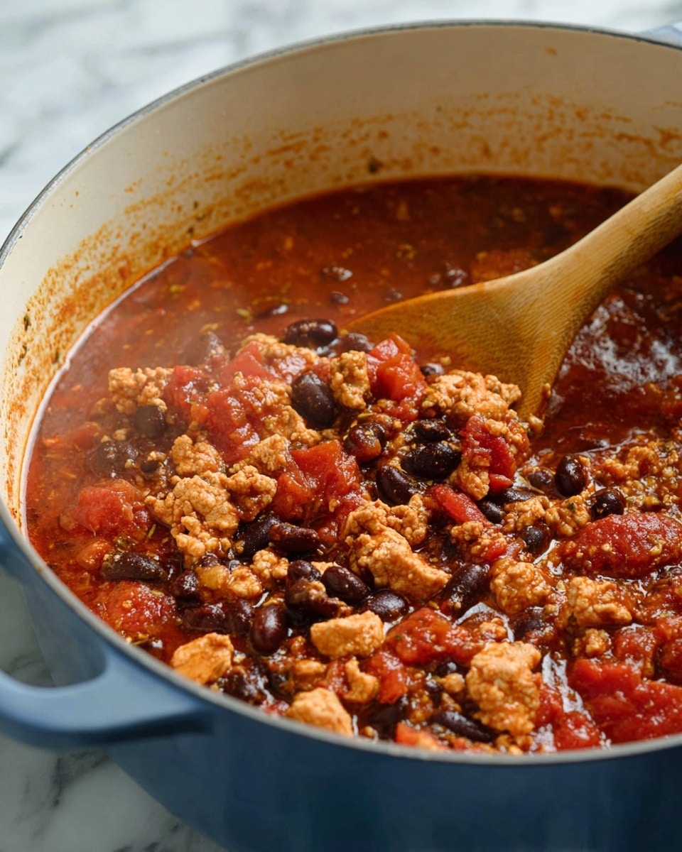 A white bowl filled with a thick reddish-brown chili soup sits on a white marbled surface. The chili has visible layers of diced tomatoes, black beans, bits of brownish ground meat, and small pieces of onion, all mixed in a rich tomato-based broth. Fresh green herbs are sprinkled on top, adding a touch of bright color. A silver spoon rests inside the bowl, partly submerged in the chili. Behind the bowl, there are green lime wedges and a white bowl with fresh green leaves. A dark blue cloth with white pinstripes lies to the left side on the white marbled surface. photo taken with an iphone --ar 4:5 --v 7