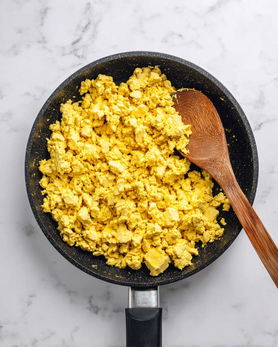 A top view of a black frying pan filled with scrambled tofu, showing a crumbly texture in bright yellow and light beige colors mixed evenly throughout. A wooden spoon is resting inside the pan to the right side, partially covered by the tofu. The pan has a black handle extending out and sits on a white marbled surface. photo taken with an iphone --ar 4:5 --v 7