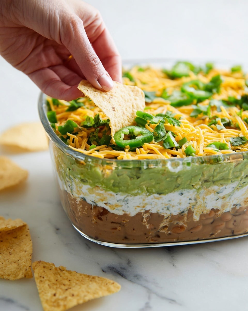 A clear glass dish shows a layered dip on a white marbled surface. At the bottom is a thick layer of refried beans. Above that is a smooth dark green layer of guacamole. Next comes a thick layer of white sour cream, then a thin dark seasoning layer on top. Above this is a thick layer of shredded yellow cheese. The top layer is decorated with green sliced scallions and fresh cilantro leaves scattered evenly. In the background, there are cut limes and a rust-colored cloth, and a few tortilla chips are on the white marbled surface nearby. photo taken with an iphone --ar 4:5 --v 7