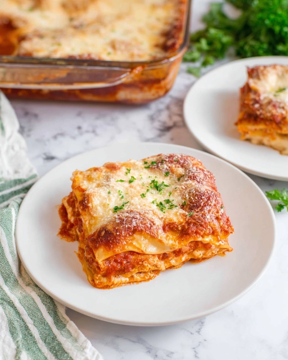 A square piece of lasagna sits on a white plate, showing around four layers: a bottom layer of red tomato sauce, a second layer with noodles and sauce mixed, a third layer of creamy cheese, and a top layer thick with melted golden-brown cheese sprinkled with grated cheese. Behind it, there is a white plate with another piece of lasagna, and at the top left, a clear rectangular baking dish filled with more lasagna. The scene is set on a white marbled surface with a green-and-white striped cloth and some green parsley leaves on the bottom right corner. Photo taken with an iphone --ar 4:5 --v 7