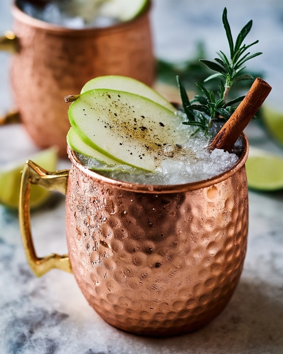 A copper cup with a hammered texture and golden handle is filled with crushed ice up to the brim. On top of the ice lies a thin green apple slice slightly angled outward, covered lightly with small black specks. A brown cinnamon stick rests on the side near the apple slice, and a small sprig of fresh green rosemary stands upright next to it. The cup sits on a white marbled surface with lime wedges visible in the blurred background. Another similar copper cup is placed behind it, partially out of focus. photo taken with an iphone --ar 4:5 --v 7