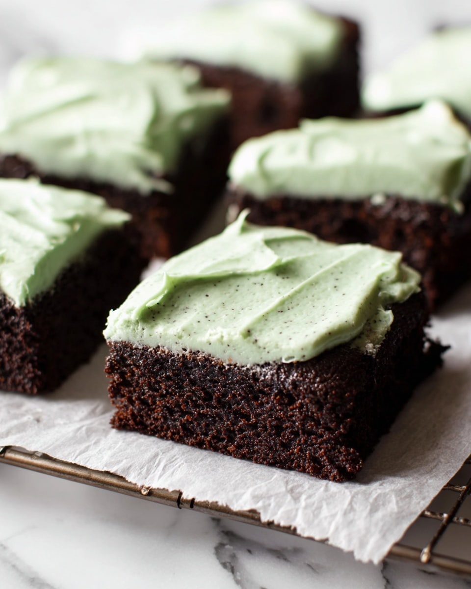 A close-up view of five square pieces of dark brown chocolate cake arranged on white parchment paper over a baking rack. Each cake piece has a thick, pale green frosting layer spread unevenly on top, showing small dark specks within the creamy texture. The cakes have a moist, dense appearance with the frosting looking smooth and rich. The background is a white marbled texture. photo taken with an iphone --ar 4:5 --v 7