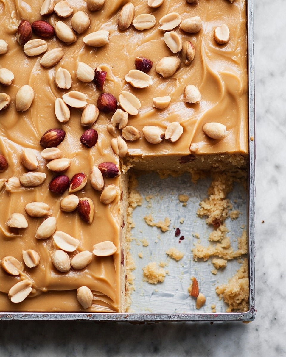 This image shows a rectangular dessert in a metal baking pan, with one piece missing from the bottom right corner. The dessert has two layers: a golden brown base layer with a slightly crumbly texture, and a top layer of smooth, light brown caramel-like frosting spread evenly. Scattered on top are whole and halved peanuts in light tan and reddish-brown colors, adding texture and color contrast. Some crumbs and small pieces of peanuts are visible near the missing piece. The pan sits on a white marbled surface. photo taken with an iphone --ar 4:5 --v 7