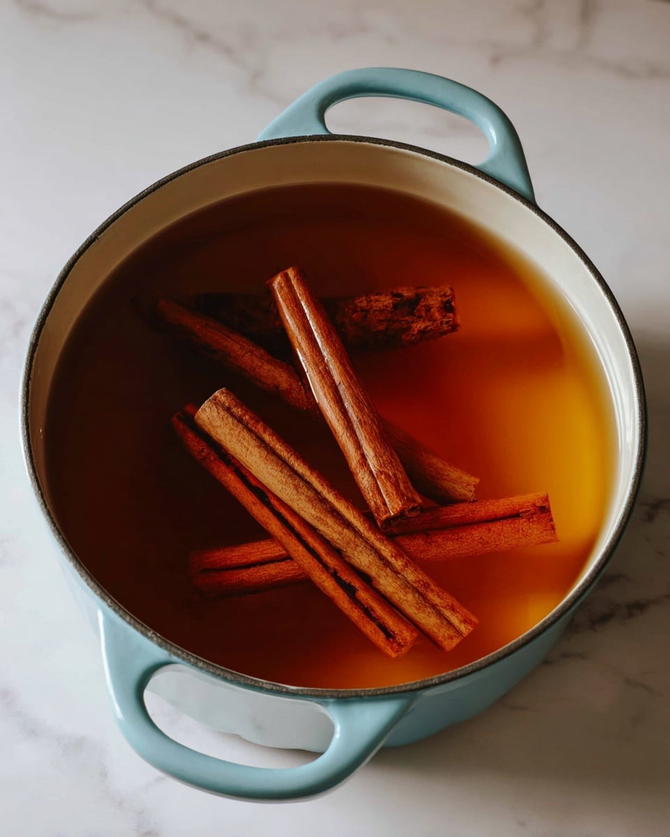 A light blue round pot with two handles holds a warm amber-brown liquid with four cinnamon sticks floating inside, varying in size and aligned across the surface; the pot is set on a white marbled surface, giving a clean and simple look. photo taken with an iphone --ar 4:5 --v 7