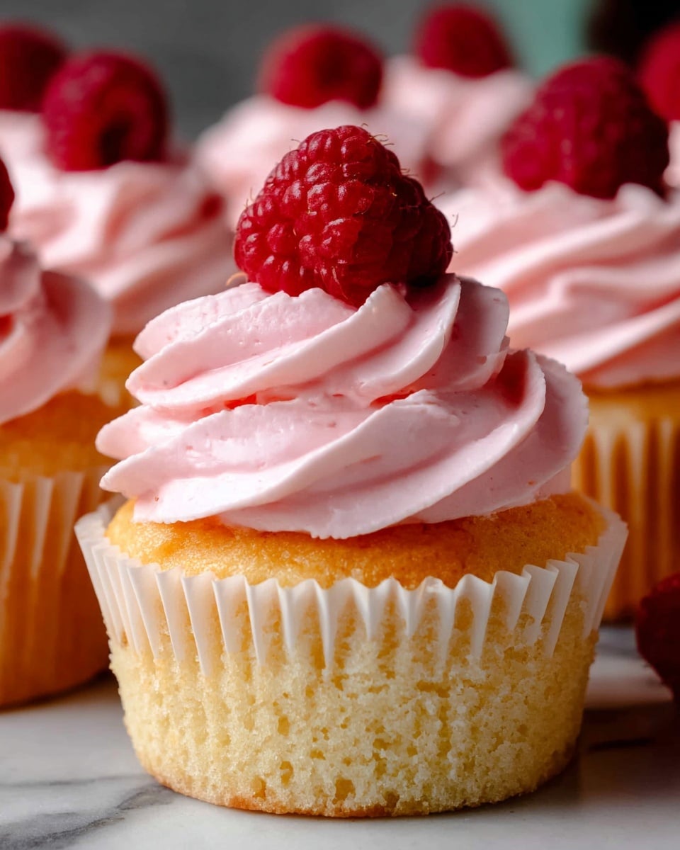 A close-up image of a cupcake with a golden yellow base wrapped in a white paper liner. On top, there is a thick swirl of light pink frosting with a smooth, creamy texture. A fresh, bright red raspberry sits on the very top center of the frosting. Surrounding the main cupcake, blurred cupcakes of the same type are visible, all set against a white marbled surface. Photo taken with an iphone --ar 4:5 --v 7