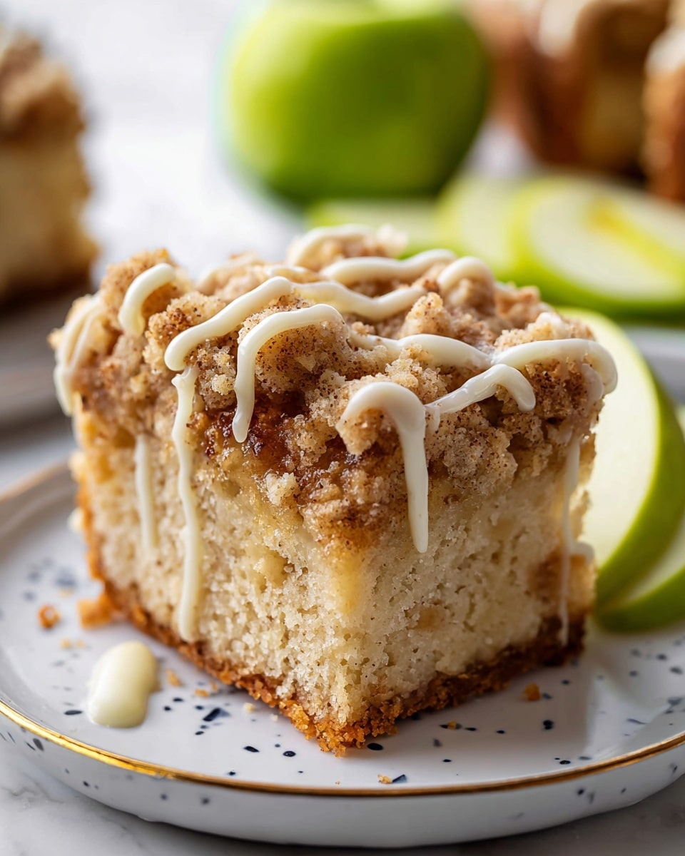 A close-up view of a crumb cake square showing three layers: a golden brown crumbly top with small lumps, a thick middle cake layer in light beige with a soft texture, and a slightly darker bottom crust. The top is drizzled with a smooth white icing in thin wavy lines. The cake is placed on a white plate with tiny blue specks and a thin gold rim, set on a white marbled surface. In the background, blurred green apple slices add a fresh color contrast. Photo taken with an iphone --ar 4:5 --v 7