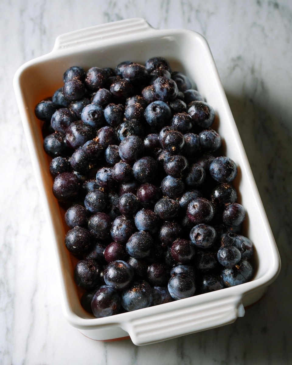 A white rectangular ceramic dish filled with a single layer of fresh blueberries evenly spread across the bottom, each blueberry having a rich dark blue and purple color with a slightly dusty texture that hints at coating or seasoning. The dish is placed on a white marbled surface, and the lighting highlights the fruit's natural gloss and round shapes. Photo taken with an iphone --ar 4:5 --v 7