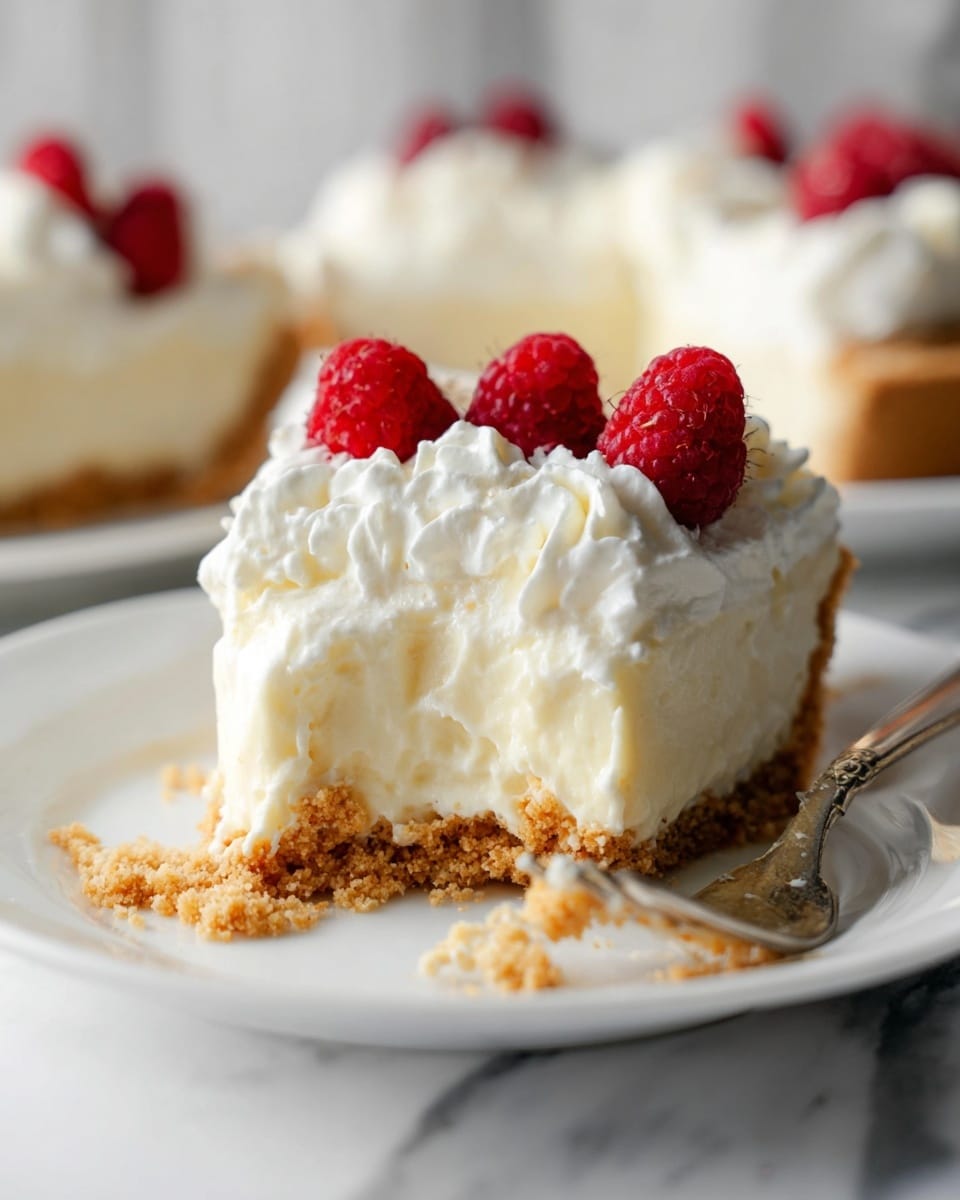 A white plate holds a slice of creamy, smooth cheesecake with three layers: a light brown crumbly biscuit base on the bottom, a thick pale white cheesecake layer in the middle, and a swirl of white whipped cream on top. Two bright red raspberries sit on the whipped cream for decoration. The plate and a silver fork rest on a white marbled surface. In the background, more slices of the same cheesecake on white plates are softly blurred, along with a small white bowl filled with raspberries. Photo taken with an iphone --ar 4:5 --v 7